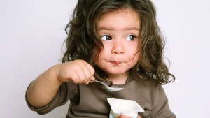 Toddler with long brown hair and brown shirt eating pudding and looking away from the camera.