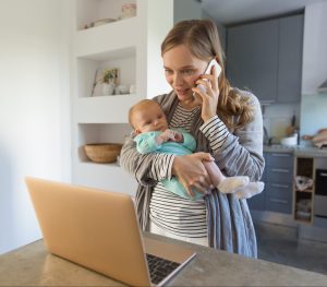 New mom holding baby and making a phone call while looking at a laptop