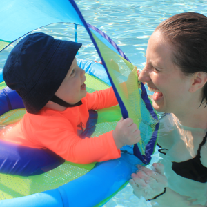 Baby in a hat and bright swim shirt smiles at mom next to him in the pool