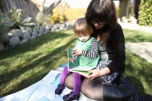 Mom and toddler read a board book together on a blanket in the shade