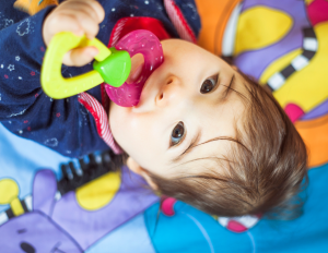Child laying on a colorful blanket looking up at the camera while chewing on a teething toy.