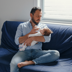 Man in blue shirt sitting on a blue couch feeding a baby a bottle