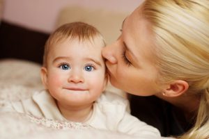 Blue eyed baby looking at the camera. Blonde mom kissing baby.