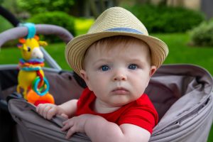 Милый 8-месячный мальчик в причудливой шляпе-федоре сидит и смотрит в камеру сбоку от своей коляски во время летней прогулки.