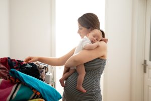 Young mother holding her baby on her shoulder is sorting laundry