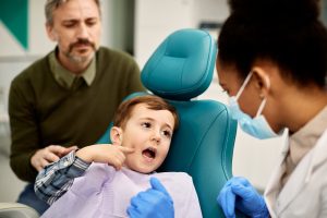 Boy in a dentist chair points to the pain in his mouth while talking to a black, female dentist.