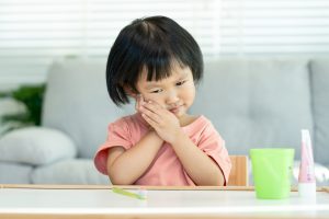 little asian girl presses hand to cheek, suffers from pain in tooth. 