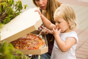 Mom and daughter open a box of pizza. A little girl licks her finger.