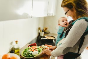 Smiling baby ported in baby carrier backpack looking at his mother while she cooks.