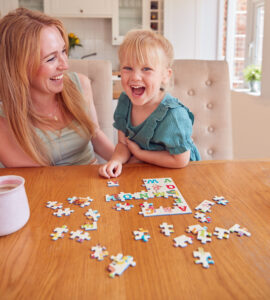 Mother And Daughter Having Fun Sitting At Table At Home Doing Jigsaw Puzzle Together