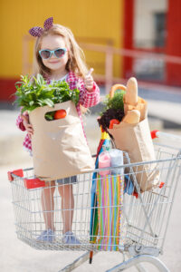 Adorable baby kid with trolley choosing fresh vegetables in local store. Girl with a big grocery cart in a parking lot near a store. A little girl with a big trolley buys fresh food and vegetables in