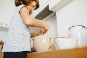 Child girl prepares a dough for baking - breaks an egg in a bowl - home cooking. Concept of food preparation, white kitchen on background.