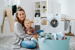 A mom sits on laundry room floor with her young daughter.