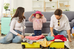Parents packing a yellow suitcase while child tries on a pink hat