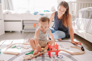 Mother and toddler boy playing with car wooden railway on floor at home. 