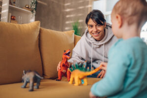 Young mother and toddler child enjoying developing activities at home while playing with dinosaurs toys on a couch. 