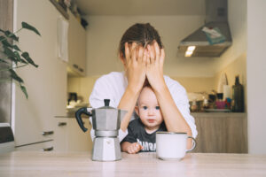 Exhausted young woman with baby is sitting with coffee in kitchen.