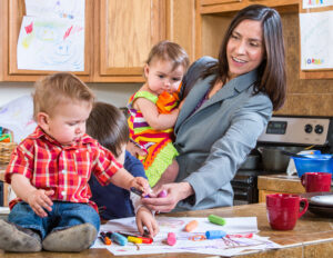Mother in business suit holding one child, while playing with two more in the kitchen.